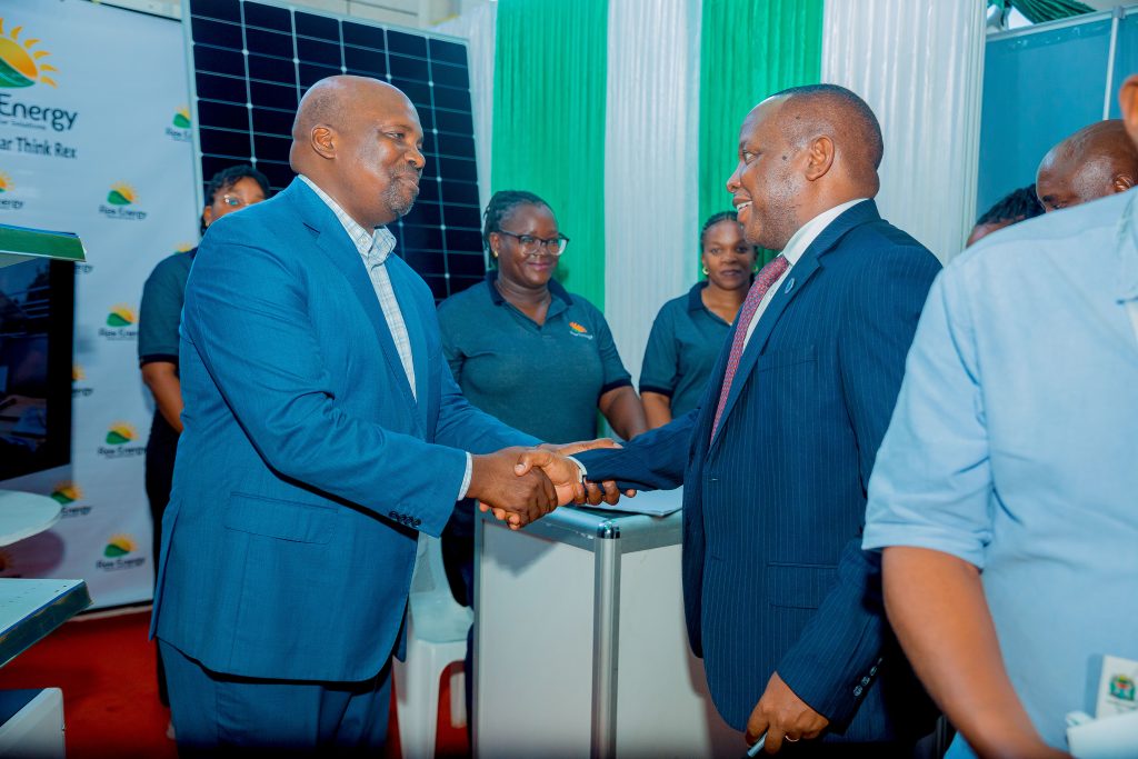 Standing in front of Rex Exhibition booth, MD Francis Kibhisa is pictures with rex employees in the background shaking hands with MoE Commisioner Eng Innocent Luoga 